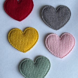 Könnte beinhalten: Fünf herzförmige Aufnäher in den Farben Rot, Grau, Gelb, Rosa und Grün. Jeder Aufnäher hat eine gestrickte Textur und einen genähten Rand. Die Herzen sind auf einem weißen Hintergrund angeordnet.
