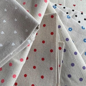 Può includere: Una collezione di campioni di tessuto piegati con una base beige naturale. I motivi includono cuori bianchi, pois rossi, pois neri, motivi floreali blu e pois viola stampati sul materiale testurizzato simile al lino.