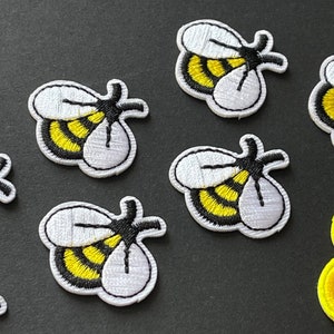 Peut inclure: Plusieurs écussons brodés en forme d'abeilles, avec des corps rayés jaunes et noirs, des ailes blanches et des contours noirs. Ils sont disposés sur une surface sombre, avec un écusson de fleur jaune et rouge. Idéal pour le bricolage.