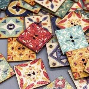 Op de afbeelding: Een verzameling vierkante houten knopen met decoratieve, kleurrijke patronen. De knopen hebben bloemen- en geometrische ontwerpen in tinten rood, blauw, geel en groen. Elke knoop heeft twee gaten om op te naaien.