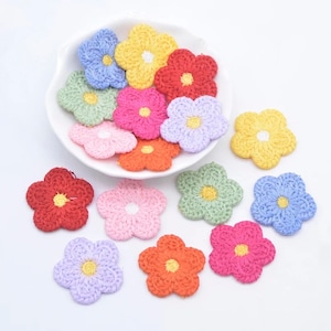 Puede incluir: Una colección de pequeñas flores de ganchillo de cinco pétalos en varios colores, incluyendo rojo, amarillo, azul, rosa, naranja, verde y lavanda. Algunas flores están dispuestas dentro de un cuenco blanco festoneado y otras esparcidas.