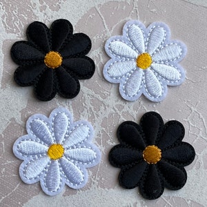 May include: Four daisy-shaped patches. Two are black with yellow centres, and two are white with yellow centres and white stitching. These embroidered patches are suitable for various craft projects.