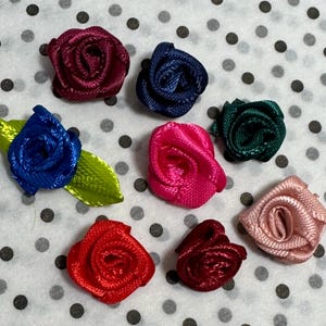 May include: A collection of eight small fabric ribbon roses in various colors including deep red, navy blue, bright blue with green leaves, hot pink, dark green, light pink, bright red, and maroon, arranged in a scattered pattern.