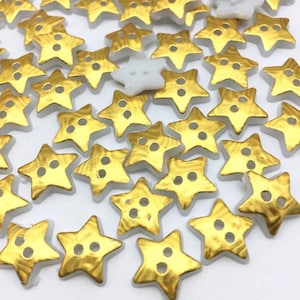 Könnte beinhalten: Eine Sammlung sternförmiger Knöpfe mit goldfarbener Metallic-Oberfläche. Jeder Knopf hat zwei Löcher zum Annähen. Ein weißer sternförmiger Knopf ist ebenfalls zu sehen. Ideal für Näh- und Bastelprojekte.