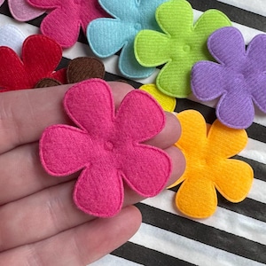 Può includere: Una collezione di abbellimenti floreali in feltro colorato in varie tonalità, tra cui rosa, viola, verde e giallo. I fiori sono disposti su una superficie a strisce bianche e nere, mettendo in risalto i loro colori vivaci e il design semplice.