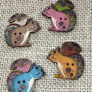 May include: Four wooden squirrel buttons in brown, pink, blue, and orange. Each button has a white belly, a patterned tail, and a floral design. The buttons are displayed on a textured, neutral-coloured surface.