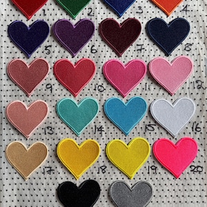 Può includere: Una collezione di ventidue toppe ricamate a forma di cuore in vari colori, tra cui rosso, verde, blu, arancione, viola, rosa e giallo. Ogni toppa ha un bordo cucito ed è esposta su un tessuto bianco con piccoli pois neri.