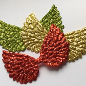 May include: Four glittery angel wings in shades of green, gold, and red-orange. The wings are arranged on a white surface, with the red-orange pair in the foreground. Each wing has a detailed, textured design.