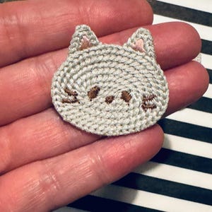 May include: A light gray embroidered cat face patch with brown details for the eyes and nose. The cat's ears have pink inner ear details. The patch is round and has a textured appearance.