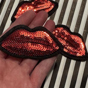 Puede incluir: Tres parches de labios rojos con lentejuelas y bordes de fieltro negro. Las lentejuelas brillan. Los parches están dispuestos sobre un fondo a rayas blancas y negras, lo que sugiere un tema artesanal o de moda.