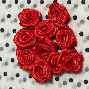May include: A collection of eight small, vibrant red ribbon roses arranged in a cluster. Each rose is crafted from tightly wound satin ribbon, featuring a spiral centre and layered petals that create a dimensional, textured floral appearance.