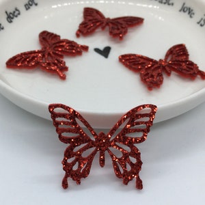 Può includere: Decorazioni a farfalla rosse glitterate. Sono mostrate quattro farfalle decorative, una in primo piano e tre su un piatto di ceramica bianco. Le farfalle hanno intricati disegni ritagliati e sono ricoperte di glitter rossi.