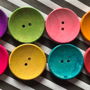 Può includere: Otto bottoni rotondi in legno di vari colori: viola, verde, rosa, arancione, rosso, giallo, turchese e fucsia. Ogni bottone ha due fori per cucire. I bottoni sono disposti su uno sfondo a righe bianche e nere.