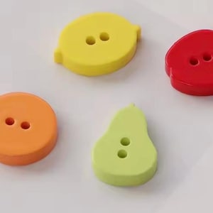 May include: Four fruit-shaped buttons with two holes each. The set includes a yellow lemon-shaped button, a red apple-shaped button, an orange circular button, and a light green pear-shaped button. Each button has a smooth, solid-coloured finish.