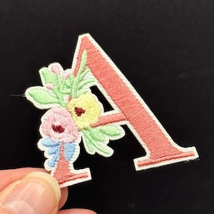 Op de afbeelding: Geborduurde patch in de vorm van de letter "A" in koraal met een bloemenontwerp. De bloemen hebben roze, gele en blauwe accenten met groene bladeren. De patch staat op een zwarte achtergrond.