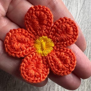 May include: A close-up of a handmade orange crochet flower with a yellow centre. The flower has five petals and is held in a hand. The crochet work is detailed and textured, showcasing the craftsmanship.