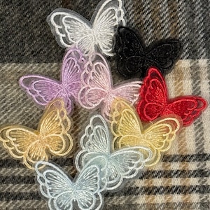 Può includere: Una collezione di applicazioni a farfalla ricamate in vari colori, tra cui bianco, nero, rosso, lavanda e oro. Le farfalle sono realizzate in tessuto trasparente con cuciture dettagliate, disposte su uno sfondo di tessuto fantasia.