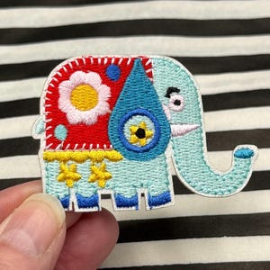 Può includere: Toppa ricamata a forma di elefante con un design colorato. L'elefante è azzurro con una sezione rossa e blu con un fiore bianco e dettagli gialli. La toppa è tenuta su uno sfondo a strisce bianche e nere.