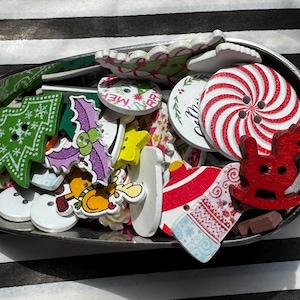 Puede incluir: Una colección de botones de madera con temática navideña en una cuchara plateada. Los botones presentan varios diseños festivos, incluyendo un árbol de Navidad, un copo de nieve y un remolino de bastón de caramelo. Los colores incluyen rojo, verde, blanco y morado.