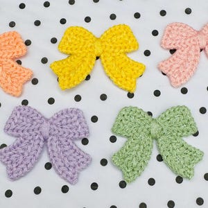 May include: Five handmade crochet bows in various colors: peach, yellow, pink, lavender, and green. The bows are arranged on a white background with black polka dots. Each bow has a textured, knitted appearance.
