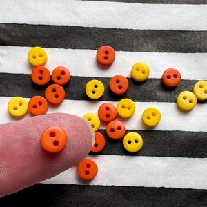Könnte beinhalten: Eine Sammlung kleiner, runder Knöpfe in Orange- und Gelbtönen. Jeder Knopf hat zwei Löcher und ist auf einem schwarz-weiß gestreiften Hintergrund verstreut. Ein oranger Knopf wird von einem Finger gehalten.