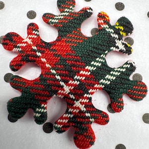 Puede incluir: Adorno de copo de nieve de tela con un patrón de cuadros rojos, verdes, negros y blancos. El copo de nieve está sobre un fondo blanco con lunares negros. El adorno tiene seis puntas.