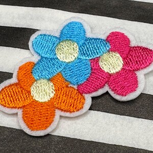 May include: Embroidered flower patch with three flowers in orange, blue, and pink. Each flower has a yellow centre and a white border. The patch is set against a black and white striped background.