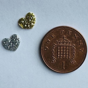 May include: Two heart-shaped embellishments, one silver and one gold, next to a penny. The hearts are covered in glitter. The penny is copper-colored and shows the number 1 and the words "ONE PENNY".
