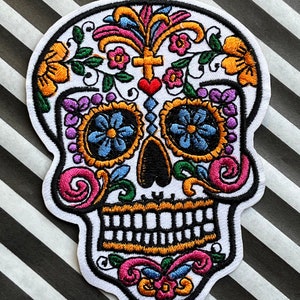May include: Embroidered patch of a sugar skull design, with a white base and colourful floral and decorative elements. The design includes orange, pink, blue, and purple accents, with a black outline. The patch is set against a black and white striped background.