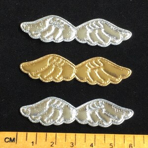 May include: Three embroidered wing patches in silver and gold. The patches are arranged on a black surface. A ruler is at the bottom of the image, with measurements in centimetres.