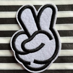 Pode incluir: Um emblema bordado branco em forma de mão de desenho animado fazendo um sinal de paz. O design apresenta contornos pretos grossos bordados que definem os dedos e a palma da mão, criando uma aparência gráfica ousada sobre a base branca.