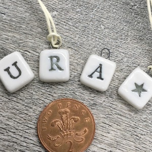 May include: Four white square pendants with the letters "URA" and a star. Each pendant is attached to a cream cord. A two pence coin is in the foreground.