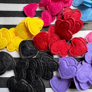Può includere: Una collezione di cuori in feltro di vari colori, tra cui rosa, rosso, giallo, nero e viola. I cuori sono sparsi su uno sfondo a righe bianche e nere, creando una composizione visivamente accattivante.