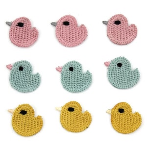 Könnte beinhalten: Neun gestrickte Entenaufnäher in Rosa, Blau und Gelb. Jede Ente hat ein schwarzes Auge und einen kleinen Schnabel. Die Aufnäher sind in einem Raster von drei mal drei auf weißem Hintergrund angeordnet. Ideal für Bastel- und Nähprojekte.
