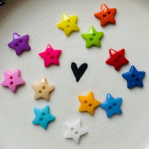 May include: A selection of star-shaped buttons in a range of colours: purple, pink, yellow, green, orange, red, blue, and white. Each button has two holes for sewing. A black heart is drawn in the centre.