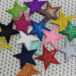 Könnte beinhalten: Eine Sammlung glitzernder sternförmiger Aufnäher in verschiedenen Farben, darunter Grün, Lila, Braun, Blau, Rosa und Silber. Jeder Stern hat einen kontrastierenden Rand und ist auf einem gepunkteten Hintergrund angeordnet. Geeignet für Bastelarbeiten und Verzierungen.