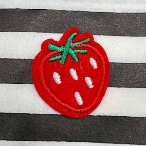 May include: A red strawberry patch with green leaves and white seeds. The patch is embroidered with a red outline and is set against a black and white striped background.