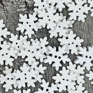 Op de afbeelding: Een verzameling witte houten knopen in de vorm van sneeuwvlokken. Elke knoop heeft zes punten en drie kleine gaatjes om op te naaien. De knopen liggen verspreid op een getextureerd, grijs oppervlak.
