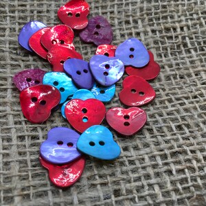 May include: A selection of heart-shaped buttons in red, purple, and blue hues. Each button features two sewing holes. The buttons are arranged on a textured, tan fabric background.