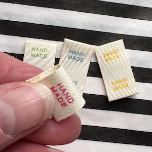 Puede incluir: Primer plano de varias etiquetas de tela rectangulares pequeñas con las palabras "HAND MADE" impresas en varios colores. Las etiquetas son de color crema con el texto en verde, azul, amarillo y rosa. El fondo es de rayas blancas y negras.
