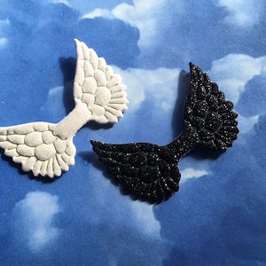 Può includere: Due decorazioni a forma di ala. Una è bianca con superficie strutturata, l'altra nera con glitter. Le ali sono poste su uno sfondo blu con un motivo a cielo nuvoloso. Ideali per lavori creativi o decorazioni.