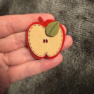 May include: A red apple-shaped felt applique with a tan centre, green leaf, and red stem. The applique has a stitched outline and two small black button details. A measuring tape is in the bottom right corner.