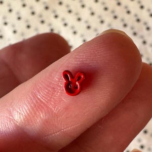 May include: A small, red button in the shape of a bunny's head. The button has two small holes for sewing and is resting on a fingertip. The button is a bright, solid red color.