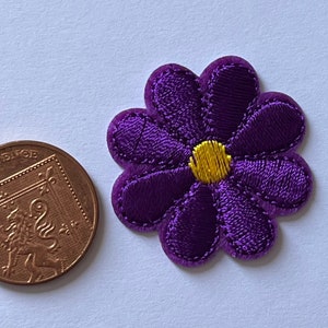 Może przedstawiać: Fioletowa, haftowana naszywka w kształcie kwiatu z ż&oacute;łtym środkiem. Naszywka ma średnicę około 3,2 cm. Do por&oacute;wnania rozmiaru dołączono brytyjską monetę o nominale dw&oacute;ch pens&oacute;w.