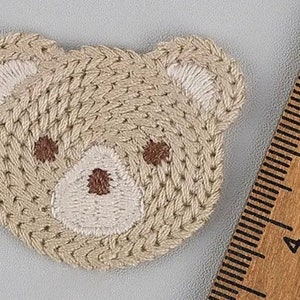 May include: Embroidered patch of a teddy bear's head. The patch has a beige woven texture with white and brown embroidered details for the ears, eyes, and nose. A wooden ruler is in the background.