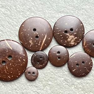 May include: A collection of seven round, dark brown buttons of varying sizes, each featuring two central holes. The buttons display a natural, mottled texture with lighter brown flecks and streaks across their smooth, polished surfaces.