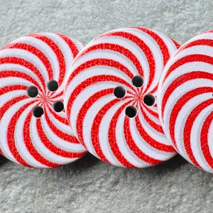 Può includere: Quattro bottoni rotondi con un design a spirale rosso e bianco. Ogni bottone ha quattro fori neri per cucire. I bottoni sono disposti in fila su una superficie grigia.