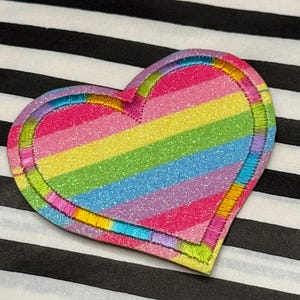 May include: A glittery, rainbow-striped heart-shaped patch with a stitched border. The patch features vibrant colours including pink, yellow, green, blue, and purple. The border is outlined with a multi-coloured stitch.