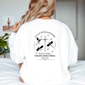 May include: White sweatshirt with a black graphic print of a sword and three dragons. The text reads "You look all frail and breakable. But you're really a violent little thing, aren't you?"