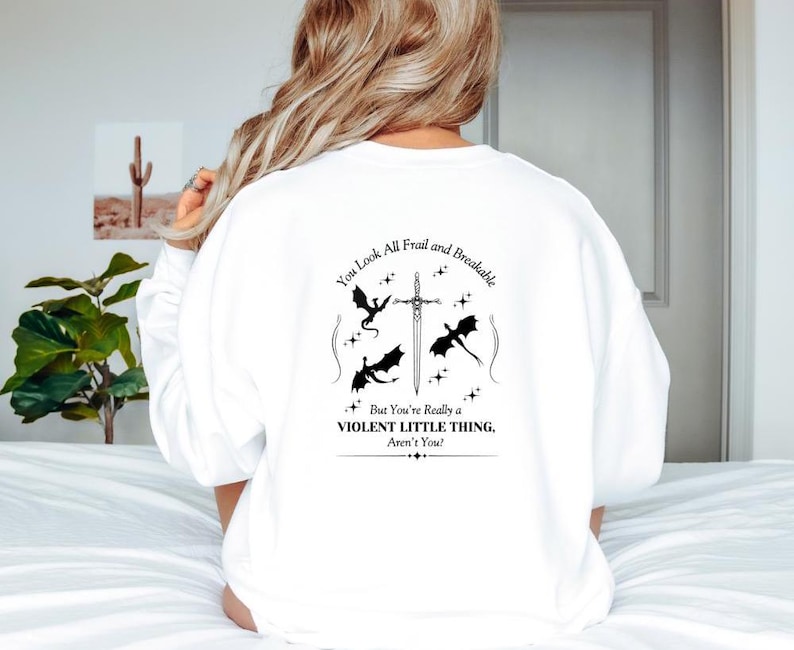May include: White sweatshirt with a black graphic print of a sword and three dragons. The text reads "You look all frail and breakable. But you're really a violent little thing, aren't you?"
